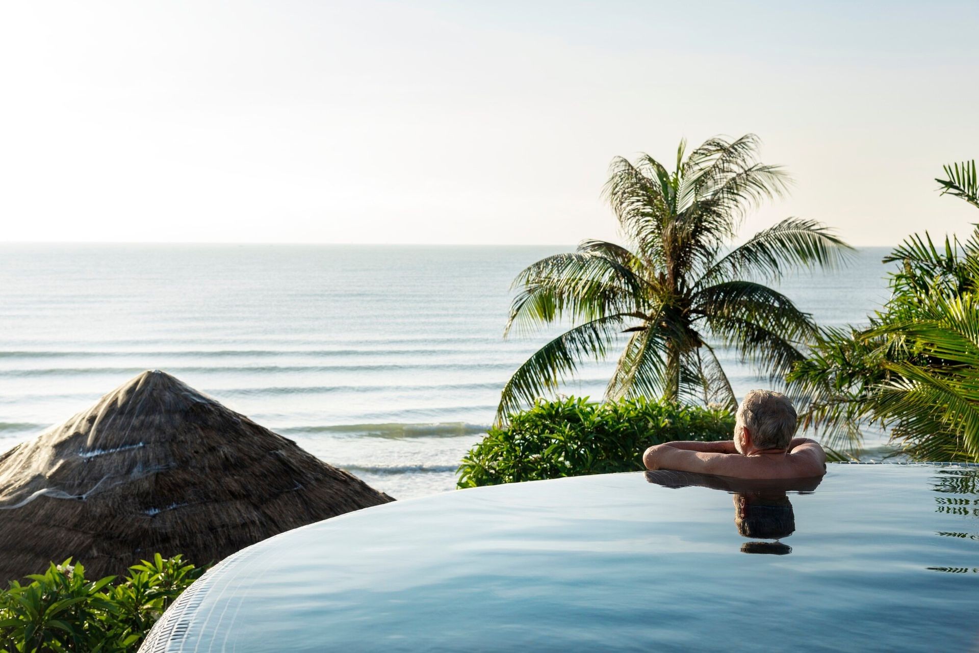 Person relaxing in an infinity pool overlooking the ocean with palm trees and a thatched roof nearby.